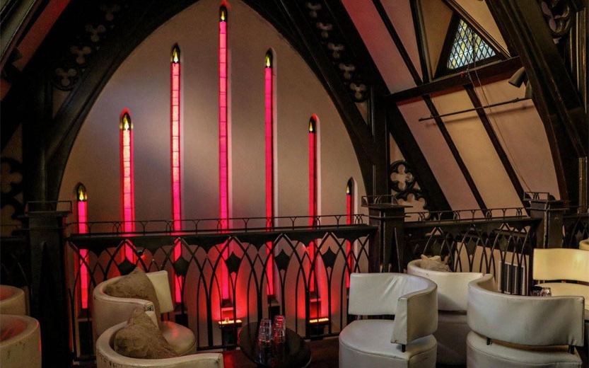 A balcony lounge with white armchairs below tall red-lit gothic panels.