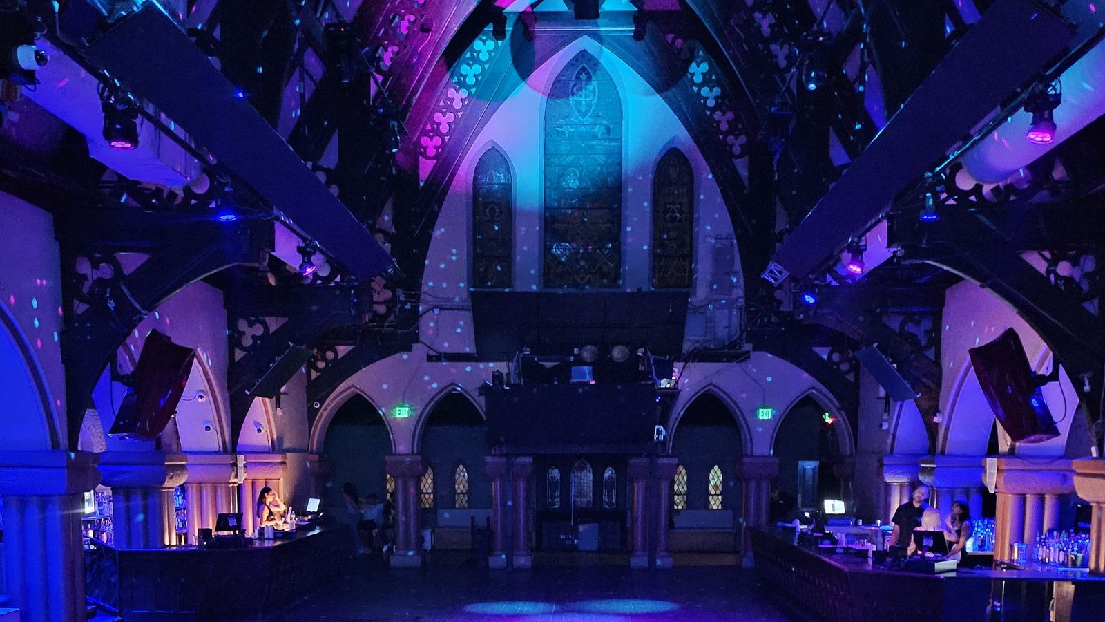 The full length of The Church bathed in purple light, gothic arches and a stained glass window at the far end.